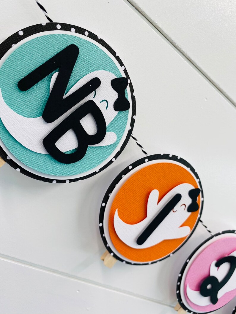 May include: A Halloween banner featuring three round paper circles. Each circle displays a white ghost with a black bow and a black letter on a coloured background. The first circle has a teal background with the letter "N", the second circle has an orange background with the letter "B", and the third circle has a pink background with the letter "1".