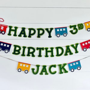 May include: A green and white Happy Birthday banner with a train theme. The banner includes the words "Happy", "Birthday", "Jack", and "3rd". The banner is decorated with small red, blue, and yellow train cars.