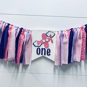 May include: A pink and blue fabric banner with the word "one" and a pink airplane with white clouds. The banner is decorated with pink, purple, and blue fabric strips.