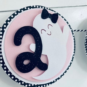K&ouml;nnte beinhalten: Ein rosa-schwarzes Halloween-Banner mit einem wei&szlig;en Geist mit einer schwarzen Schleife und der Zahl "2" in schwarzem Glitzer.