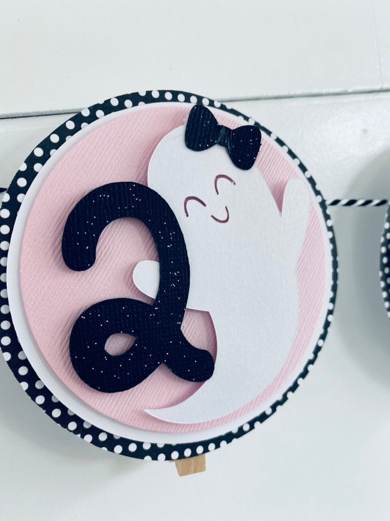 K&ouml;nnte beinhalten: Ein rosa-schwarzes Halloween-Banner mit einem wei&szlig;en Geist mit einer schwarzen Schleife und der Zahl "2" in schwarzem Glitzer.