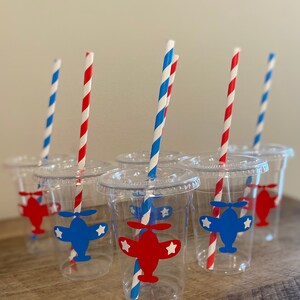 May include: Six clear plastic cups with red and blue striped straws. Each cup has a red or blue airplane sticker with white stars on it.
