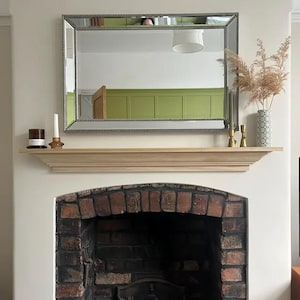 May include: A rectangular wall mirror with a beaded frame hangs above a light-coloured wooden mantelpiece. The mantel features a dark candle, a white taper candle in a glass holder, two gold-toned candle holders, and a patterned vase with dried grass.