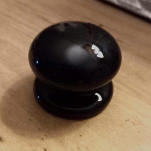 May include: A black, glossy cabinet knob with a spherical top and a flat, disc-shaped base. The knob is made of a smooth, reflective material. It is positioned on a light-coloured wooden surface.