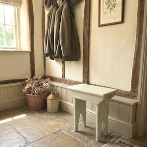 May include: A small, light-coloured wooden stool with four angled legs stands on a stone floor. The stool features a rectangular top and a simple, sturdy construction. It is positioned in a room with cream-coloured walls, wooden beams, and a woven basket.