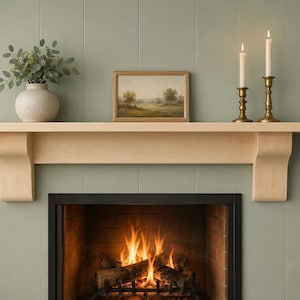 May include: A light-coloured wooden fireplace mantel shelf mounted above a fireplace with a burning fire. On the mantel sits a round ceramic vase with green foliage, a framed landscape painting, and two brass candlesticks with lit white candles.