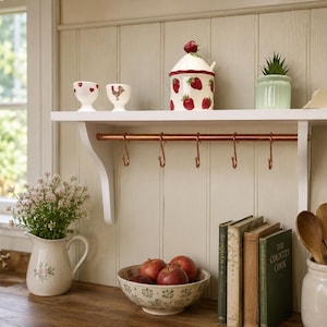 May include: A white wooden wall shelf featuring a copper rail with five S-hooks. The shelf displays two egg cups, a ceramic jar with a strawberry design and lid, a small green succulent in a ribbed pot, and a decorative cloud-shaped item.