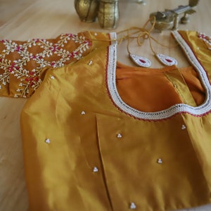 May include: A mustard yellow silk blouse with intricate floral embroidery in shades of pink, white, and gray. The neckline is adorned with a white beaded trim and a red border.
