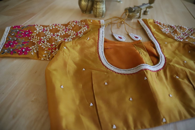 May include: A mustard yellow silk blouse with intricate floral embroidery in shades of pink, white, and gray. The neckline is adorned with a white beaded trim and a red border.