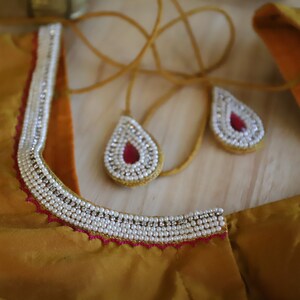 May include: A gold fabric garment with a white pearl trim and pink beaded embellishments. The garment has a neckline with a decorative beaded design.
