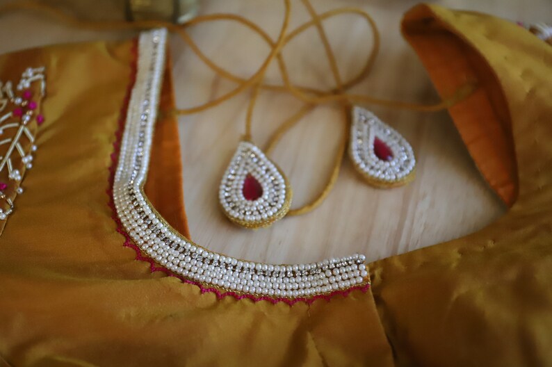 May include: A gold fabric garment with a white pearl trim and pink beaded embellishments. The garment has a neckline with a decorative beaded design.