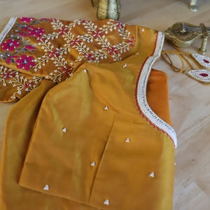 May include: A mustard yellow embroidered blouse with a white and pink floral design. The blouse has a beaded trim around the neckline.