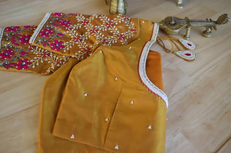 May include: A mustard yellow embroidered blouse with a white and pink floral design. The blouse has a beaded trim around the neckline.
