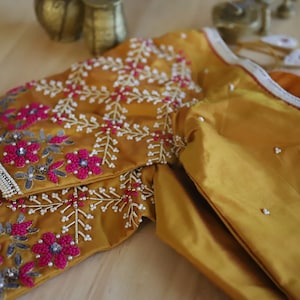 May include: A mustard yellow embroidered blouse with pink and silver embellishments. The blouse is made of a silky fabric and has a traditional Indian design.