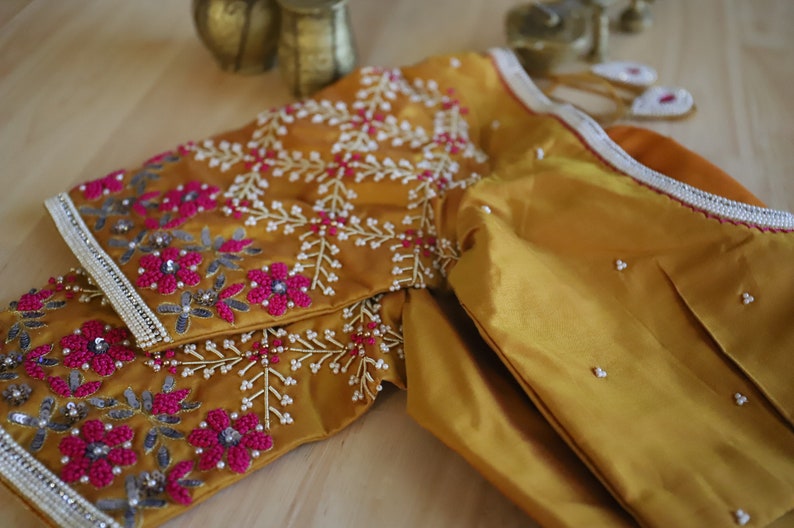 May include: A mustard yellow embroidered blouse with pink and silver embellishments. The blouse is made of a silky fabric and has a traditional Indian design.