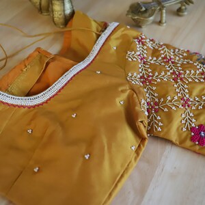 May include: A mustard yellow silk blouse with intricate embroidery featuring pink and white beads. The blouse has a beaded trim around the neckline and a beaded design on the sleeve.