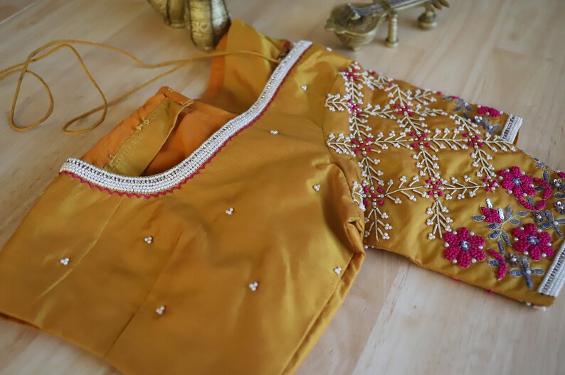 May include: A mustard yellow silk blouse with intricate embroidery featuring pink and white beads. The blouse has a beaded trim around the neckline and a beaded design on the sleeve.