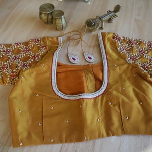 May include: A mustard yellow embroidered blouse with a round neckline and short sleeves. The blouse is decorated with white beads and pink and red floral embroidery.