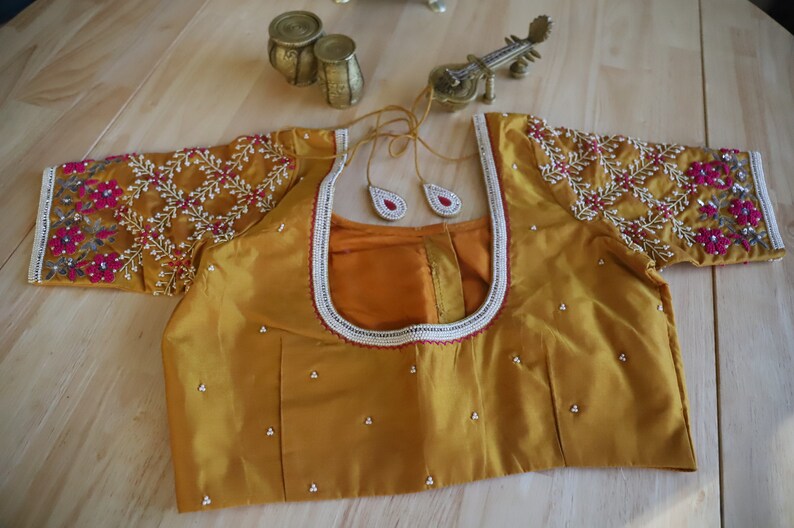 May include: A mustard yellow embroidered blouse with a round neckline and short sleeves. The blouse is decorated with white beads and pink and red floral embroidery.