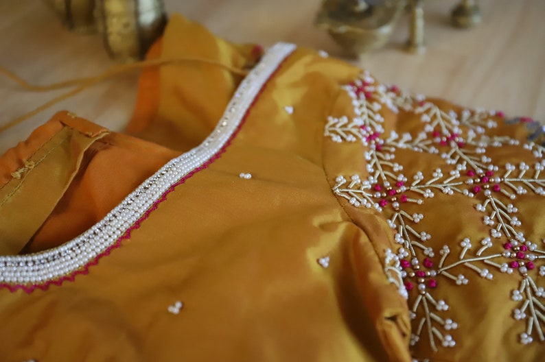 May include: A mustard yellow fabric with a white and pink beaded design. The fabric has a beaded trim around the neckline.