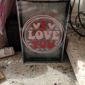 May include: A clear glass box with a black frame and a pink and red "I Love You" design with hearts.