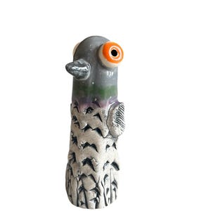 Puede incluir: Figura de paloma de cerámica con cabeza gris, ojos naranjas y cuerpo blanco y gris. El ave tiene un pico pequeño y plateado y un diseño texturizado y tallado en su cuerpo. La figura está sobre un fondo blanco.