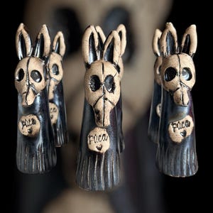 May include: A group of ceramic figurines with rabbit skull-like faces and long ears. The figurines are dark blue with tan-coloured faces and ears. Each figurine has a heart-shaped tag with the word "Puca" written on it.