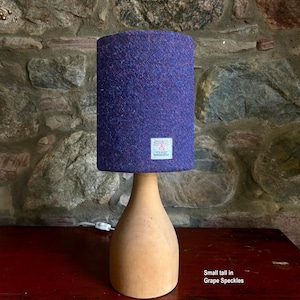 May include: A small, tall table lamp with a wooden base and a cylindrical lampshade. The lampshade is a deep purple with flecks of red and blue. The base is a light, natural wood color. The lamp is on a dark red surface.
