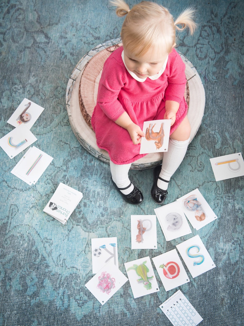 May include: A young child sits on a wooden stump, holding a flashcard with a picture of a horse. The child is wearing a pink dress and white socks. There are other flashcards scattered on the floor, each featuring a different letter of the alphabet and a corresponding picture.