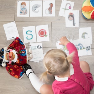 May include: A young child sits on a wooden floor surrounded by colorful alphabet flash cards. The cards feature letters, animals, and words. The child is wearing a pink shirt and has their hair in pigtails.