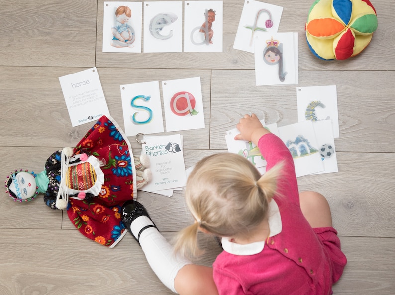 May include: A young child sits on a wooden floor surrounded by colorful alphabet flash cards. The cards feature letters, animals, and words. The child is wearing a pink shirt and has their hair in pigtails.