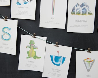 May include: A set of alphabet flashcards with illustrations and descriptions for each letter. The cards are hung on a string with clothespins. The letters include S for snake, K for kicking, L for line, M for mountains, T for t-rex, U for upside-down umbrella, and V for vase.