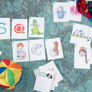 May include: A set of colorful alphabet flash cards with pictures. The cards feature a variety of illustrations, including a snake, an apple, a dinosaur, an elephant, a monkey, a mountain, a queen, a horse, and a pencil. The cards are designed to help children learn the alphabet and phonics.