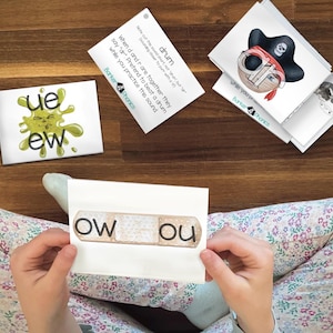 May include: Educational flashcards for phonics learning. The image shows several cards with words and illustrations, including a pirate, a green splat with the letters "ue ew", and the word "drum". A child is holding a card with the letters "ow ou".