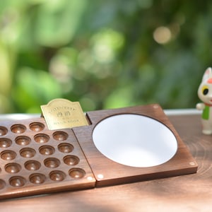 May include: A wooden organizer with a grid of small, round indentations, a mirror, and a small brass plaque with text. A small cat figurine and a cartoon cat figurine are in the background. The organizer is open.