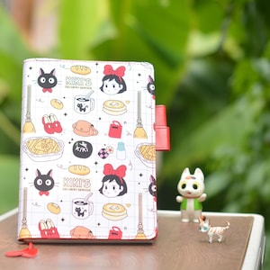 May include: A white planner with a red and black cover featuring a repeating pattern of Kiki's Delivery Service characters, including Kiki, Jiji, and various items from the movie. The planner has a red ribbon bookmark.