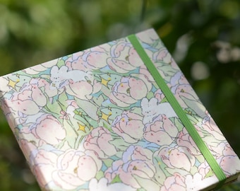May include: A small, square notebook with a green elastic band closure. The cover features a whimsical design of pink and white tulips and white bunnies against a green background.