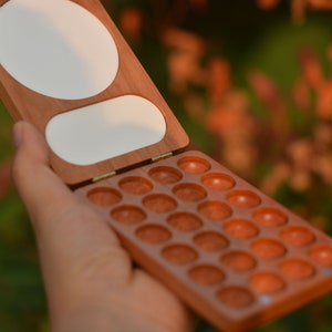 May include: Open wooden paint palette with a hinged lid. The palette is made of reddish-brown wood and contains several small, round wells for holding paint. The lid has two white, oval-shaped areas. The palette is held open, revealing the wells.
