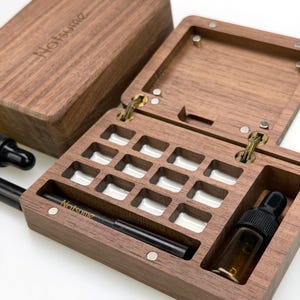 Pode incluir: Uma caixa de madeira escura com tampa articulada, aberta para revelar uma grade de pequenos compartimentos forrados de branco. A caixa é feita de madeira escura e tem o nome "Natsume" gravado na tampa e uma caneta preta. Uma pequena garrafa de vidro com conta-gotas é visível.