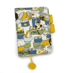 May include: A rectangular notebook cover featuring a repeating pattern of various cats sitting inside colorful metal food cans. The cover is light-colored with yellow pen loops on the right side and a yellow ribbon bookmark with two circular ends.