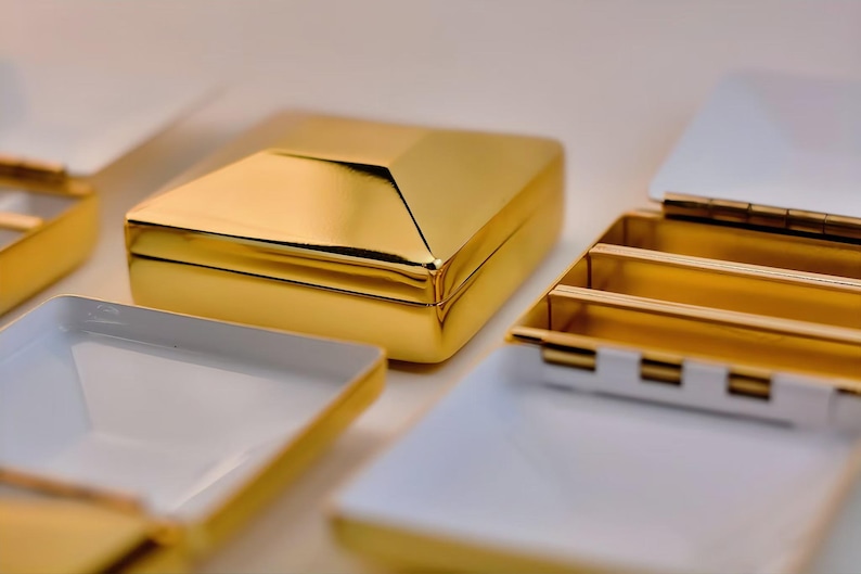 Puede incluir: Primer plano de varias cajas doradas con interiores blancos. Una caja est&aacute; cerrada, mostrando una superficie brillante y reflectante. Otras cajas est&aacute;n abiertas, revelando compartimentos y un interior blanco. Las cajas tienen una apariencia lujosa y elegante.