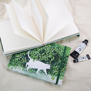 May include: Two open blank journals with a green and white cat design. The journals are open to reveal the blank pages. Two tubes of black paint are next to the journals.