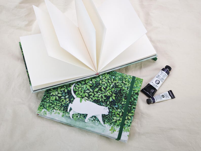 May include: Two open blank journals with a green and white cat design. The journals are open to reveal the blank pages. Two tubes of black paint are next to the journals.