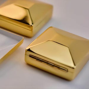 Puede incluir: Cajas geom&eacute;tricas doradas con un acabado met&aacute;lico reflectante. Una de las cajas est&aacute; abierta, revelando un interior blanco. Las cajas tienen un dise&ntilde;o moderno y angular, dispuestas sobre una superficie blanca.