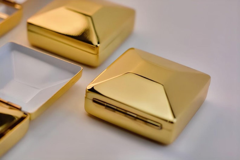 Puede incluir: Cajas geom&eacute;tricas doradas con un acabado met&aacute;lico reflectante. Una de las cajas est&aacute; abierta, revelando un interior blanco. Las cajas tienen un dise&ntilde;o moderno y angular, dispuestas sobre una superficie blanca.