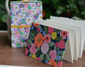 Puede incluir: Dos cuadernos con estampado floral, uno con un diseño floral rosa y morado y el otro con un diseño floral azul y naranja. Los cuadernos están cerrados y colocados sobre una superficie de madera.