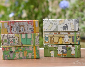 May include: A stack of four colorful memo pads featuring different illustrations of cats. Each pad has a green elastic band and a yellow tab. The cats are depicted in various poses and activities, including riding a train, eating at a restaurant, and relaxing on a couch.