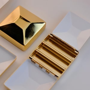 Puede incluir: Cajas de joyer&iacute;a cuadradas doradas con interiores y tapas blancas. Una caja est&aacute; cerrada, mientras que otras est&aacute;n abiertas, revelando compartimentos. Las cajas tienen un dise&ntilde;o geom&eacute;trico y una superficie brillante y reflectante. Las cajas est&aacute;n dispuestas sobre una superficie blanca.