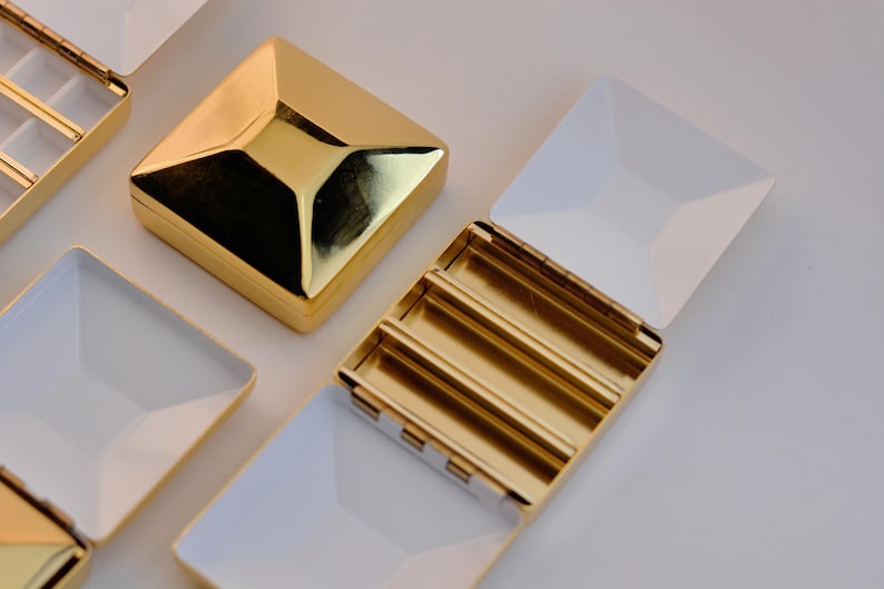 Puede incluir: Cajas de joyer&iacute;a cuadradas doradas con interiores y tapas blancas. Una caja est&aacute; cerrada, mientras que otras est&aacute;n abiertas, revelando compartimentos. Las cajas tienen un dise&ntilde;o geom&eacute;trico y una superficie brillante y reflectante. Las cajas est&aacute;n dispuestas sobre una superficie blanca.