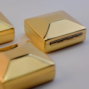 Puede incluir: Tres cajas doradas de forma cuadrada con un dise&ntilde;o geom&eacute;trico. Las cajas tienen una tapa con bisagras y una superficie brillante y reflectante. Las cajas est&aacute;n dispuestas sobre una superficie clara.